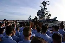 A man in a dark suit and tie speaks to sailors on an aircraft carrier. A sign in the background reads "Mission Accomplished"