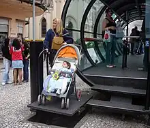 A woman with a baby carriage uses a platform lift to access a station above street level